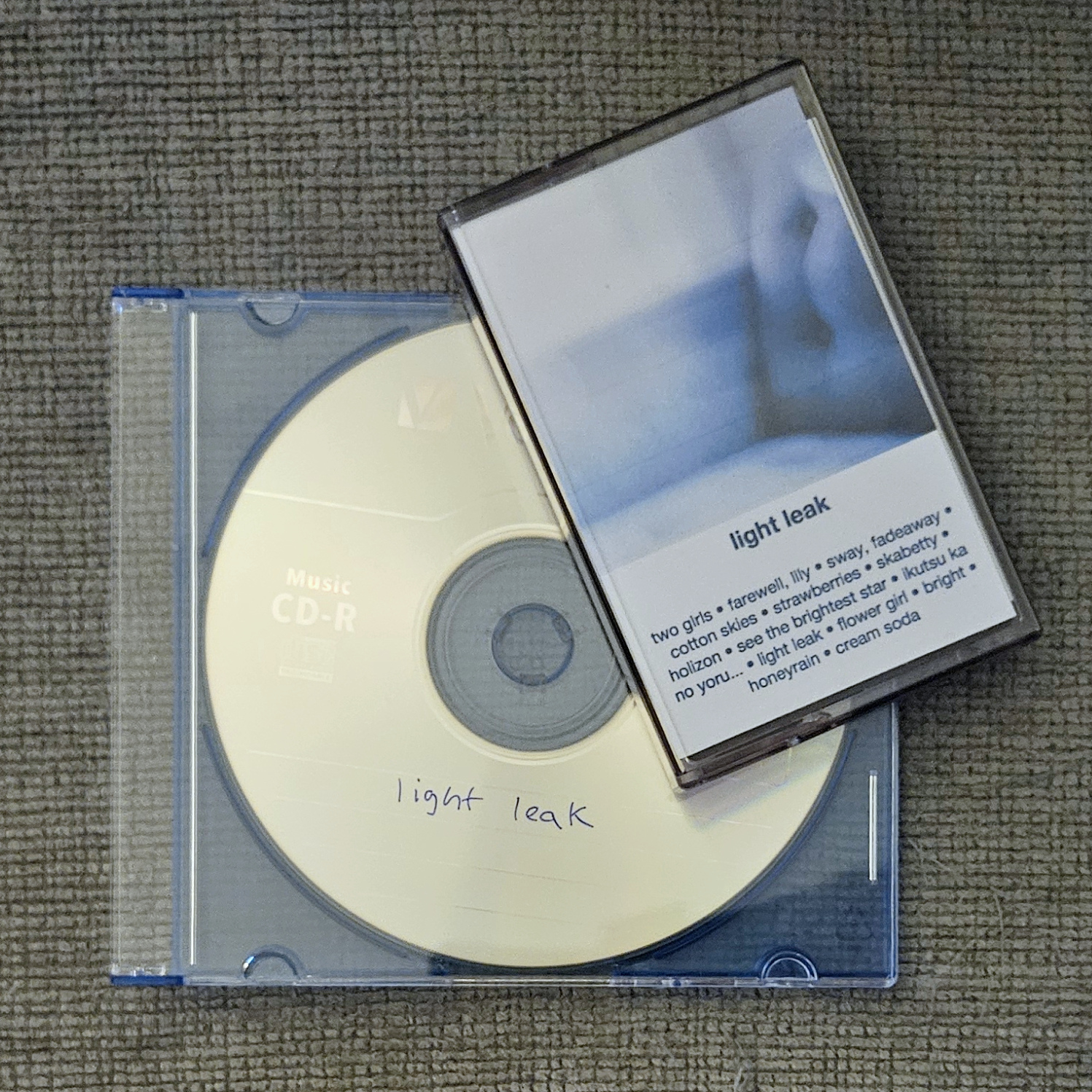 a burned CD in a translucent blue slim jewel case with a cassette tape in a case with a j-card inside on top of it diagonally. the cassette tape's j-card depicts a blue couch washed out with light leak filters and the track listing below, while the CD case has no cover and simply shows the disc inside that has 'light leak' written on it in blue ink
