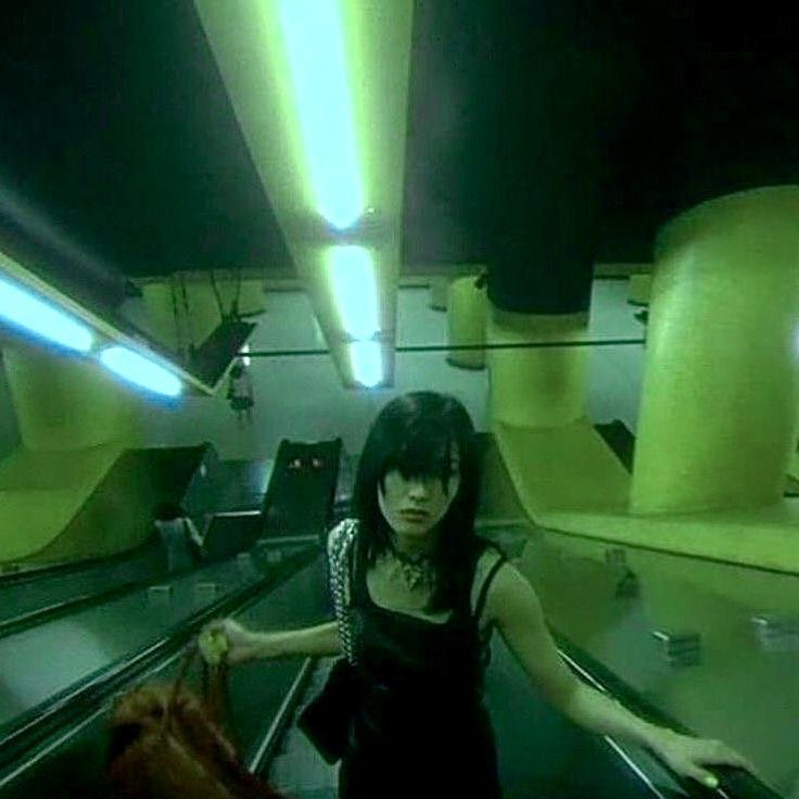 a woman on a high escalator looking solemn as she holds a bag and rests a hand on the railing. she's illuminated by vertical lamps on the ceiling and the whole image is a green hue