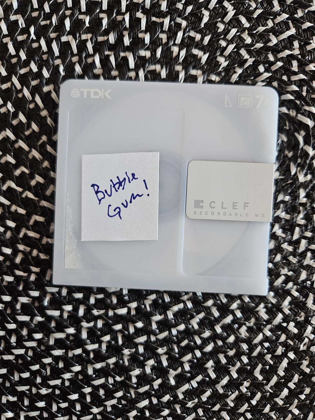 a mini-disc on a table. it has a cut label on the label section that has 'bubble gum!' written on it and the disc cartridge is an almost opaque white