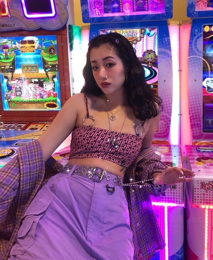 another woman in a mix of 2000s and 1980s fashion leaning on and posing in front of arcade game machines. she's also under colorful neon lighting and the game machine lights illuminate her
