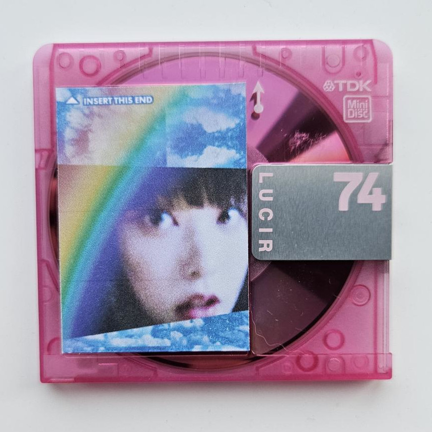 a hot pink translucent mini disc on a white sheet of paper. the mini disc has a sticker label on it that's a web-core edit of haerin from new jeans, her photo at a warped angle with pixelated distorted blue and white sparkling clouds tiled behind her and a big translucent rainbow on top of the whole image