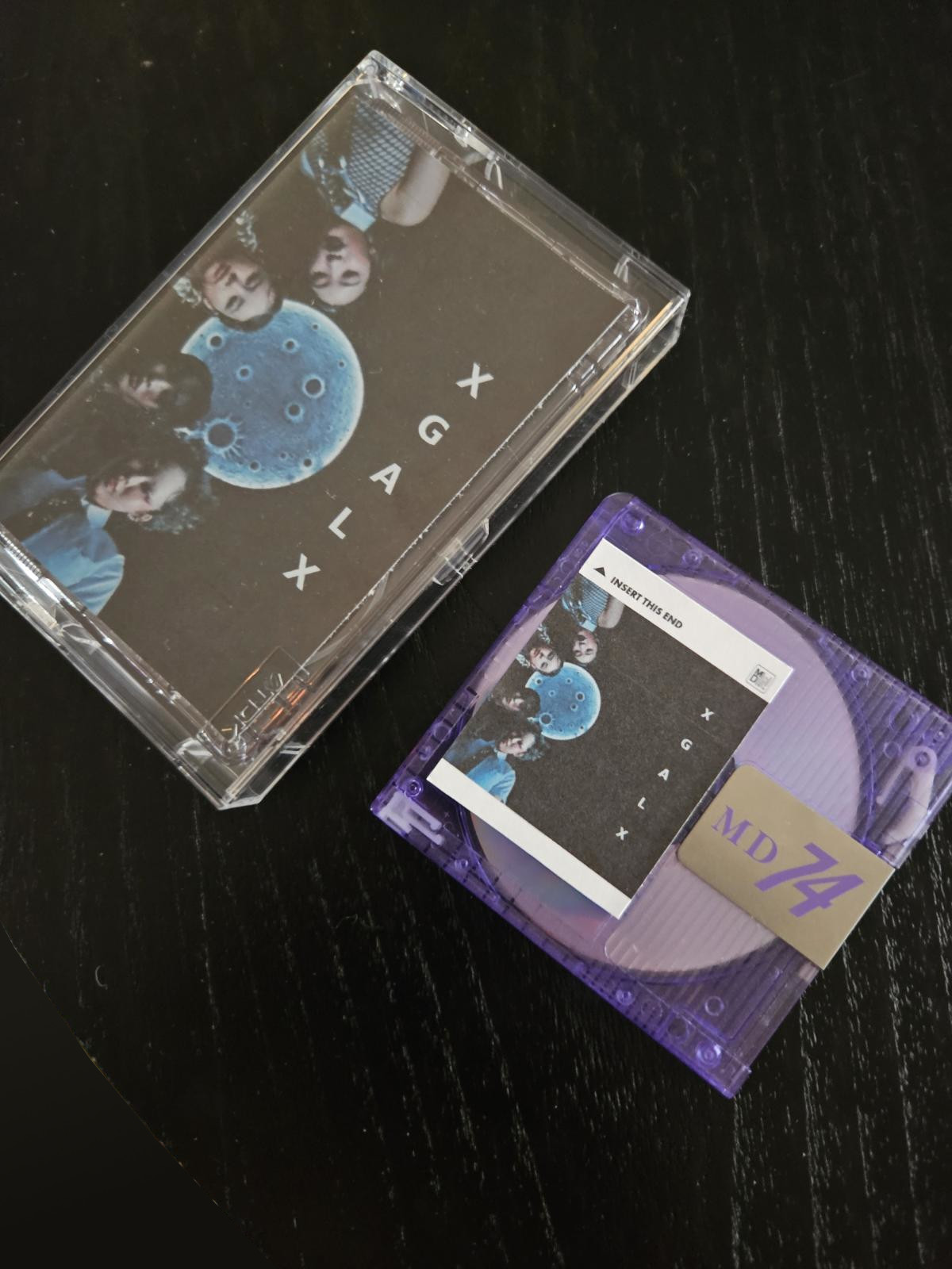 a cassette tape in a clear plastic case and a purple daiso mini disc next to it. the tape has a j-card and the mini disc a matching label, both vary but have the same image of the XG rap line members in the trampoline thumbnail with 'X G A L X' spelled out vertically beside it