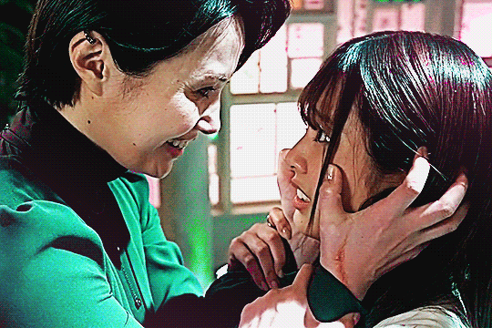 kamen rider ZEZTZ GIF showing the lady as she grips nem by the face with both hands, the lady going from smiling creepily to a stern, almost foreboding expression, as nem looks on in horror and grips the lady's arms. the colors are similar to the last gif, with cool-toned greens and cyans and magenta highlights