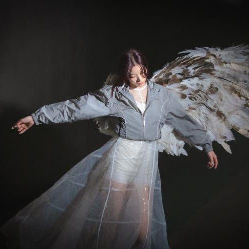 kazuha from LE SSERAFIM wearing a half zipped up jacket and a thin white long tiered skirt, with her arms out as if dancing and a burnt angel wing behind her, the other half of it burnt off