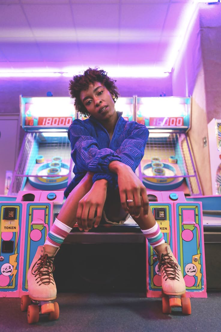 a woman sitting and leaning forward with her knees together and hands folded over them. she's wearing roller skates and a 1980s style outfit and sits in front of arcade game machines. she's under colorful neon lighting