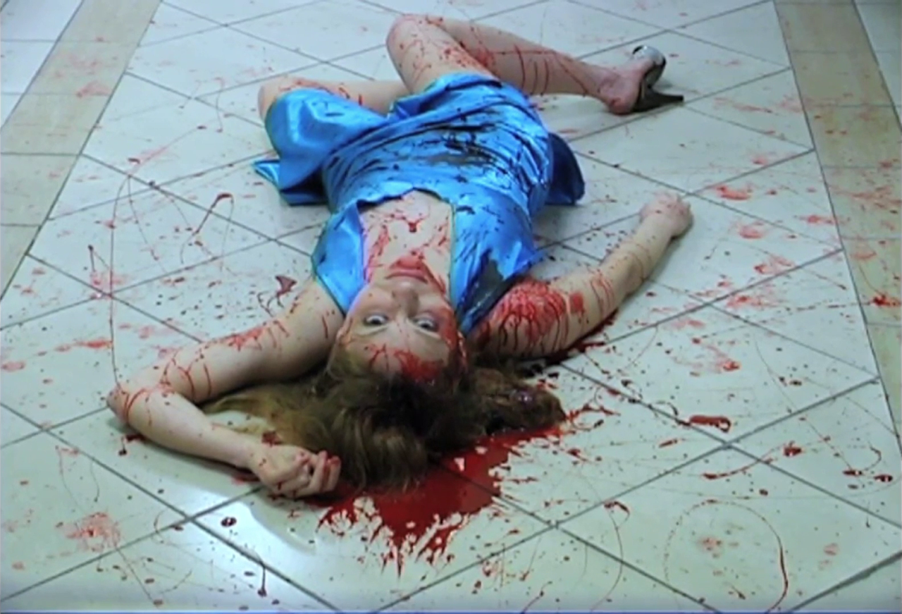 naomi elizabeth on a blood stained floor in a shiny deep blue sleeveless dress. she's covered in blood splatter and looks up at the camera which photographs her from upside down