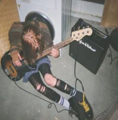 blurred photo of a person sitting on the floor with a guitar in their hands. they seem to be playing passionately with their head tipped down and towards the guitar, which is plugged into an amp beside them