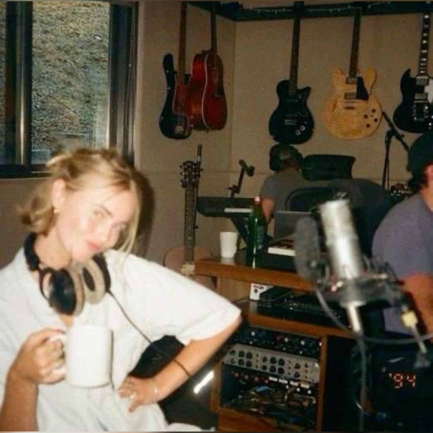 blurred photo of a girl posing and smiling with headphones around her neck in a room full of audio equipment and with guitars mounted to the walls